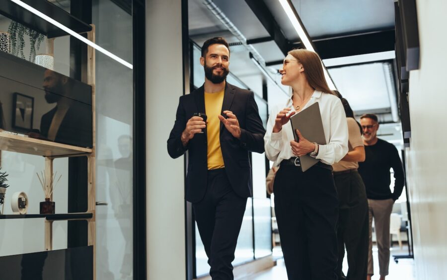 Two people in professional attire walk and converse in a modern office hallway, discussing market research. One is holding a coffee cup, while the other carries a laptop. Others are seen working in the background.